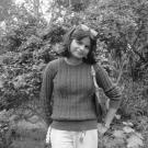 Young smiling woman in sweater standing before leafy bushes, black-and-white
